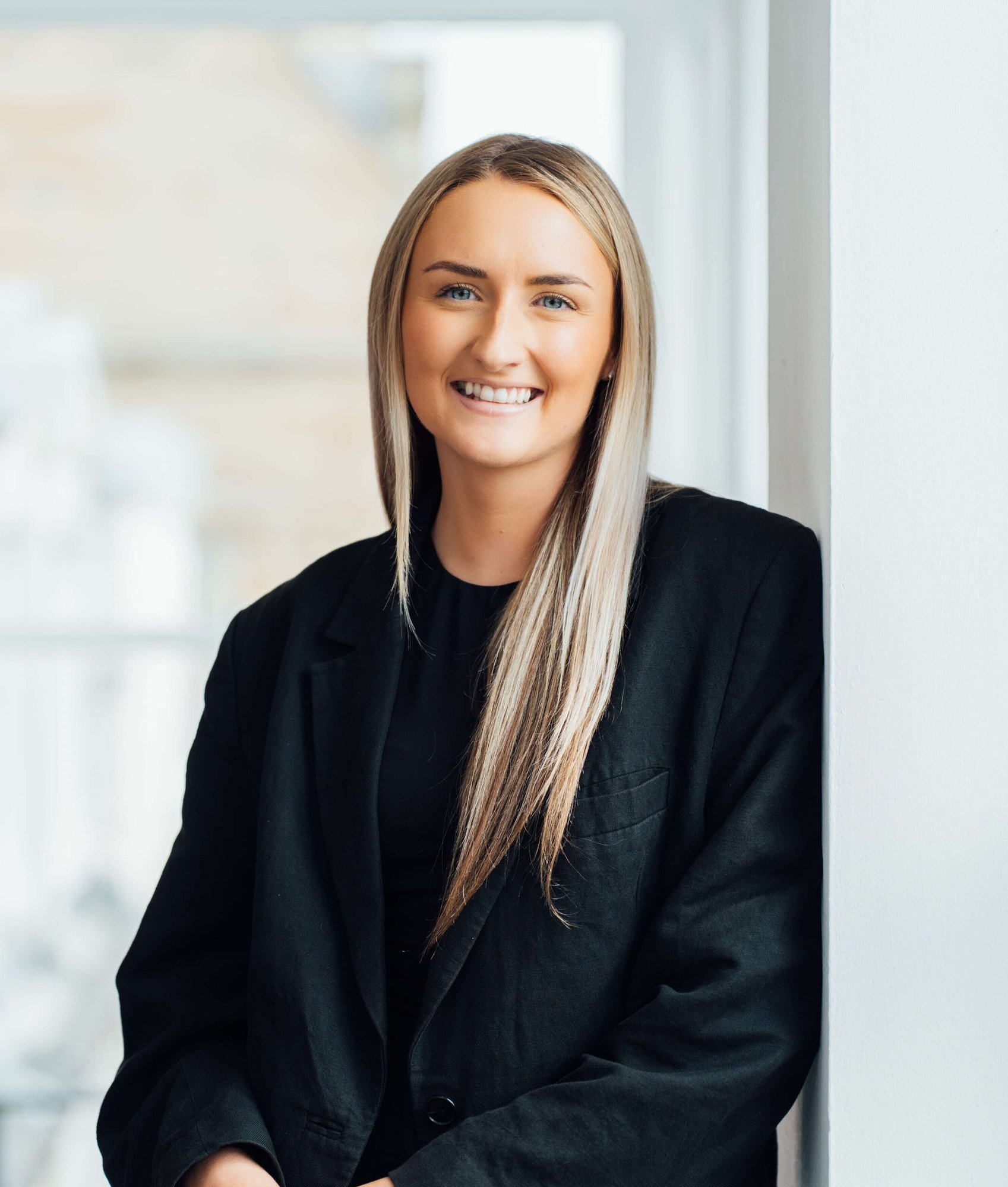 An image of Emily Ross smiling at the camera. She has blonde hair and is wearing a black blazer.