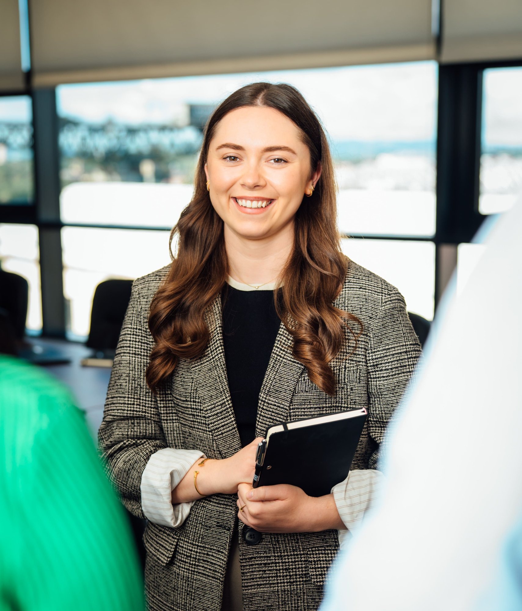 An image of Zoe Dollard smiling at the camera holding her notebook. She is wearing a black top and a brown blazer.